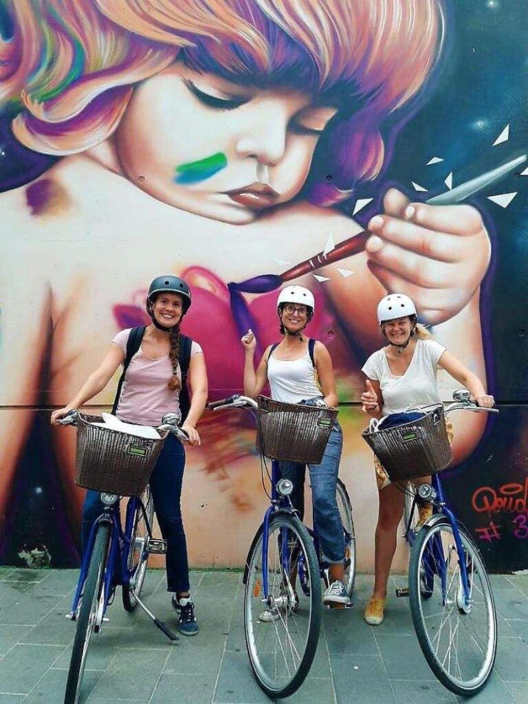 three girl tourists enjoying a bike tour and posing for a photo with a street art background