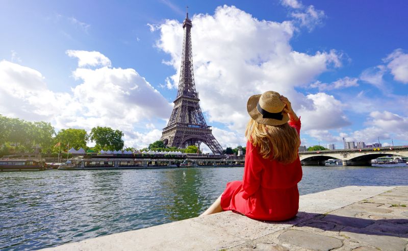 Young traveler woman in red dress and hat sitting on the quay of Seine River looking at Eiffel Tower