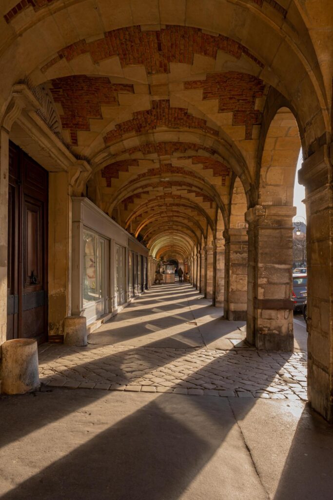 Sunlit arcade at Place des Vosges, Paris, showcasing historic architecture and cobblestone paths.