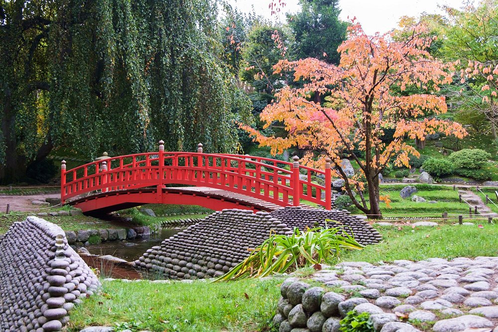 Vibrant red bridge crosses a serene stream in the park of the Albert Kahn Museum and Garden, framed by trees.