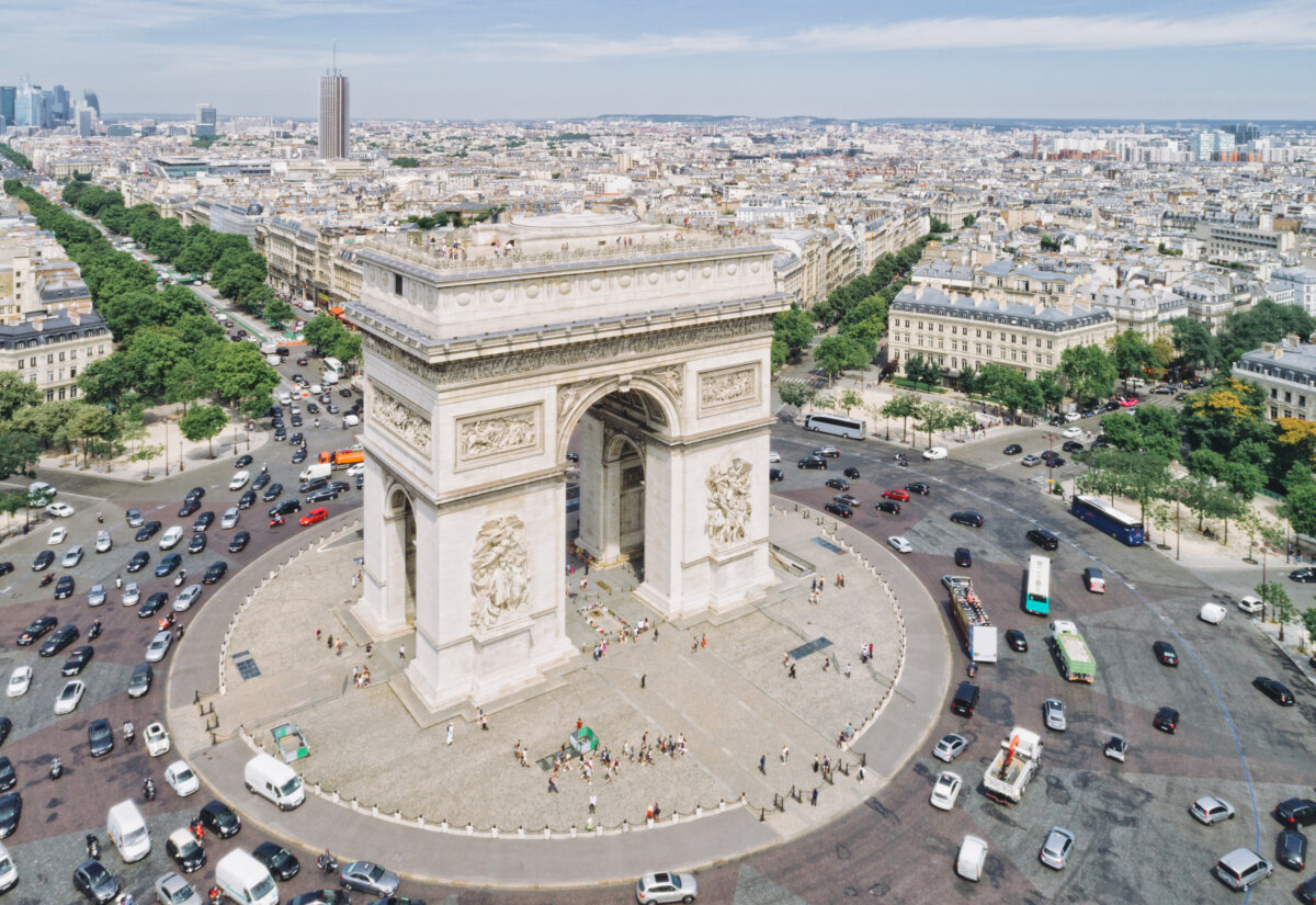 Aerial view of the Arc de Triomphe in Paris, surrounded by a circular traffic roundabout filled with vehicles. The cityscape extends into the distance.
