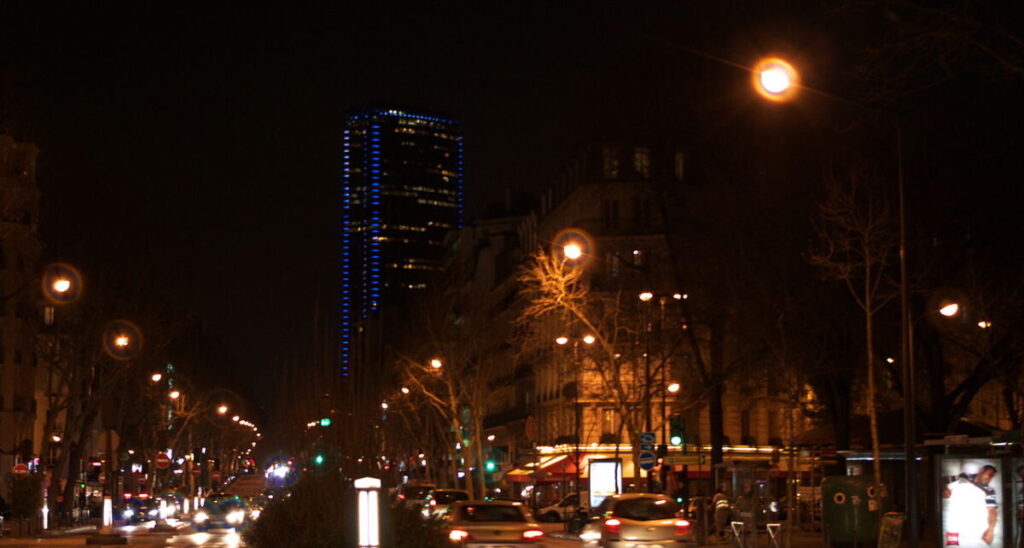 Avenue du Maine at night, featuring Montparnasse Tower, with glowing streetlights and a lively urban scene.