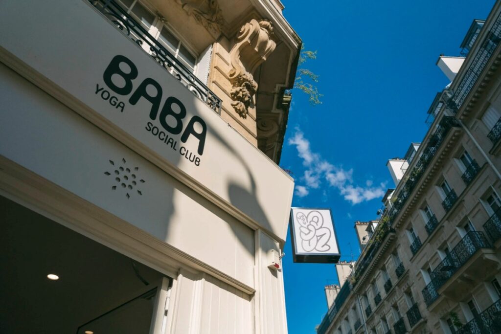 Front of "Baba Yoga Club" against a bright blue sky in a Parisian street, with ornate architecture and a playful logo on the sign.