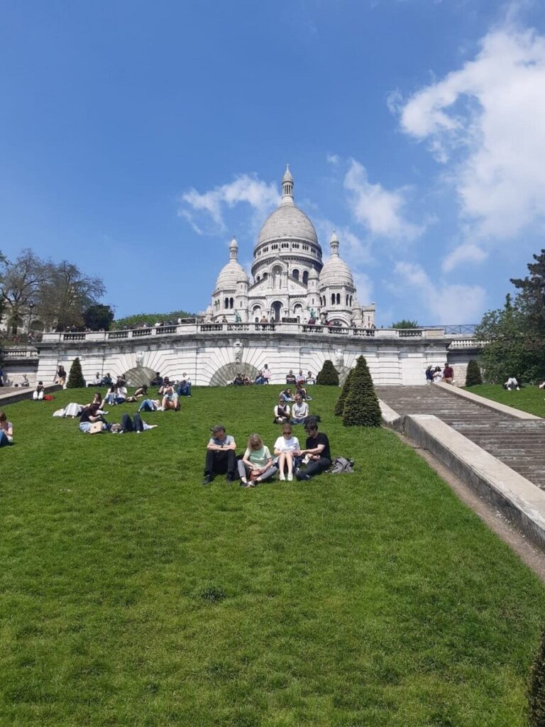 People relaxing on a grassy hill with the Sacré-Cœur Basilica in the background. The sky is clear and blue, creating a peaceful, sunny atmosphere.