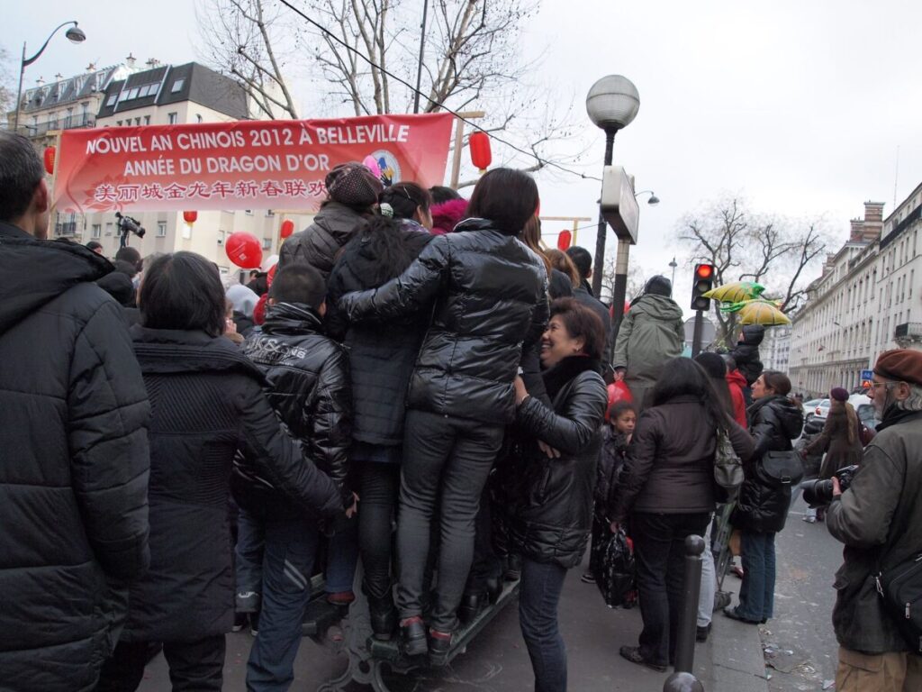 Crowd celebrating Chinese New Year 2012 in Belleville, with people in black jackets gathered under a red banner. Festive, lively atmosphere.