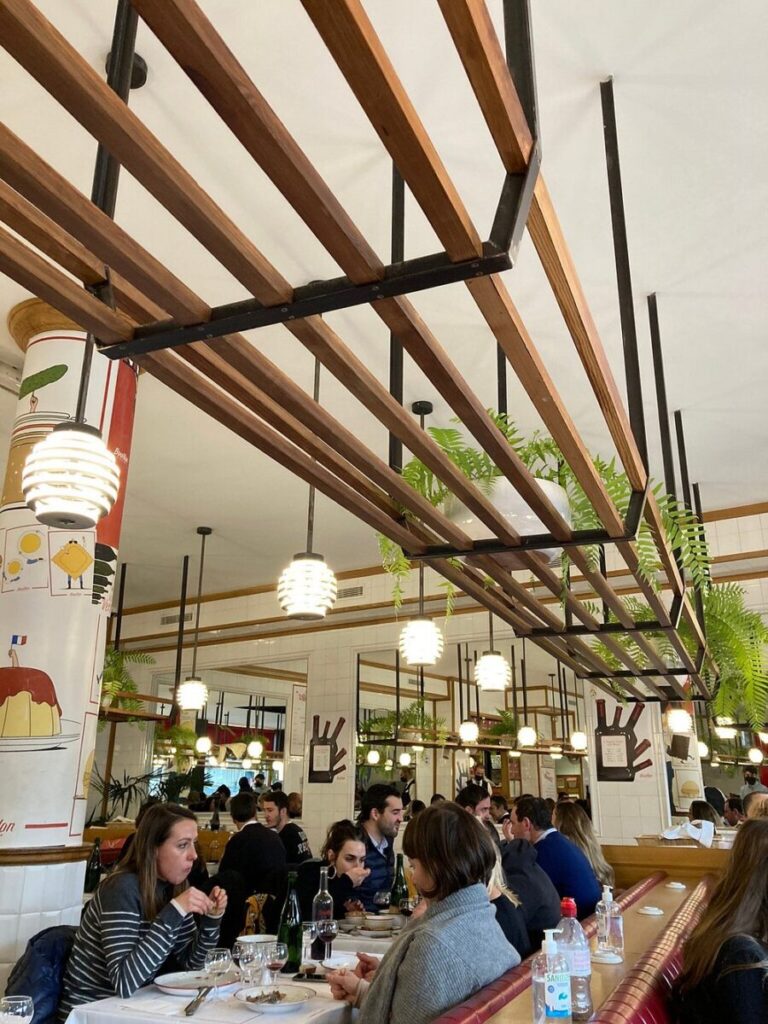 Bouillon Pigalle lively restaurant interior with diners seated at long tables under wooden ceiling beams. Hanging lights and plants create a cozy, vibrant atmosphere.