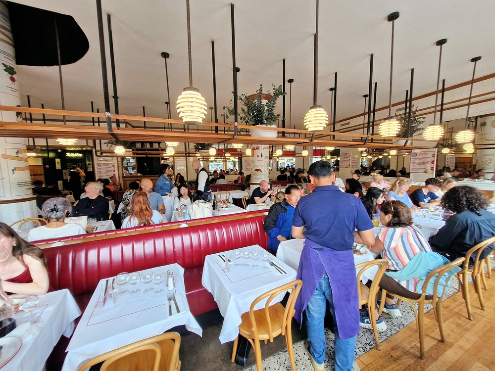 Bouillon Pigalle restaurant with wooden chairs and red booths. Diners engage in conversations under round pendant lights, creating a lively, inviting atmosphere.