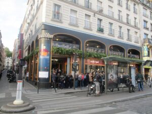 Corner view of a bustling Parisian café with large windows and greenery above. People are gathered outside near a bus stop, conveying a lively atmosphere.