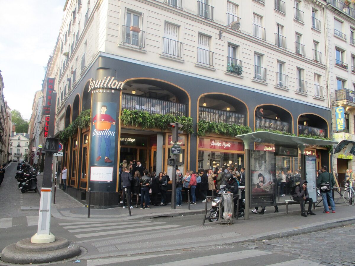 Corner view of a bustling Parisian café with large windows and greenery above. People are gathered outside near a bus stop, conveying a lively atmosphere.