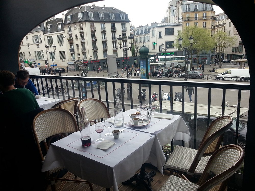 Cozy Bouillon Pigalle balcony overlooking a bustling street with elegant buildings. Tables set with wine and coffee cups. Atmosphere is lively yet relaxed.