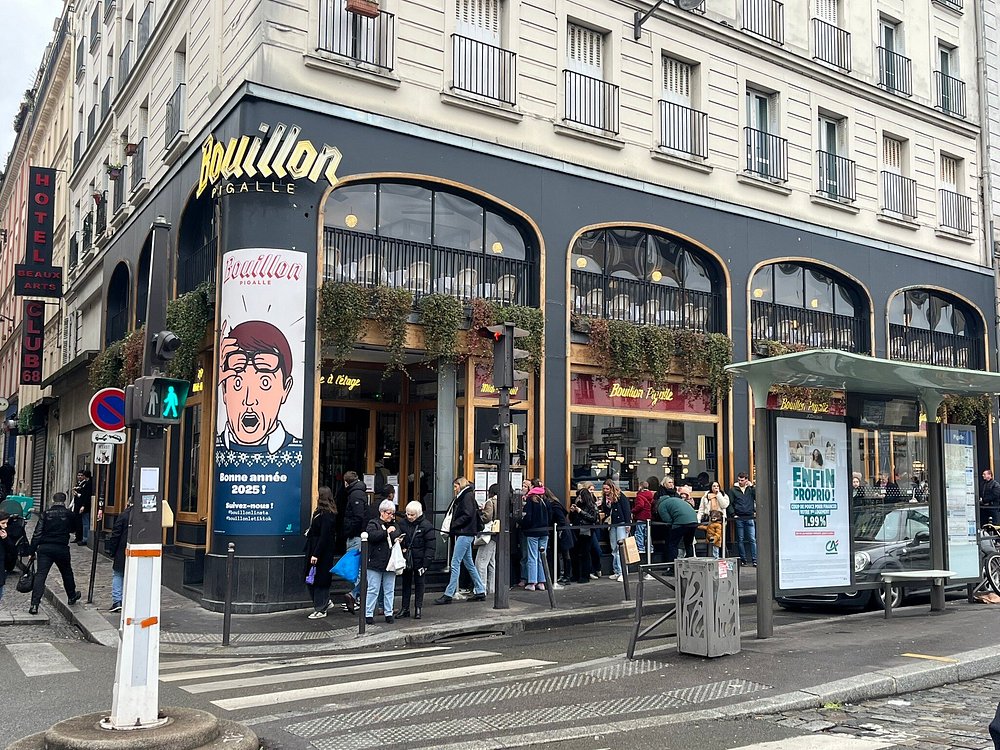 Street scene with people outside a restaurant, "Bouillon Pigalle," featuring a retro-style poster. The building has large windows and green hanging plants, conveying a lively urban atmosphere.