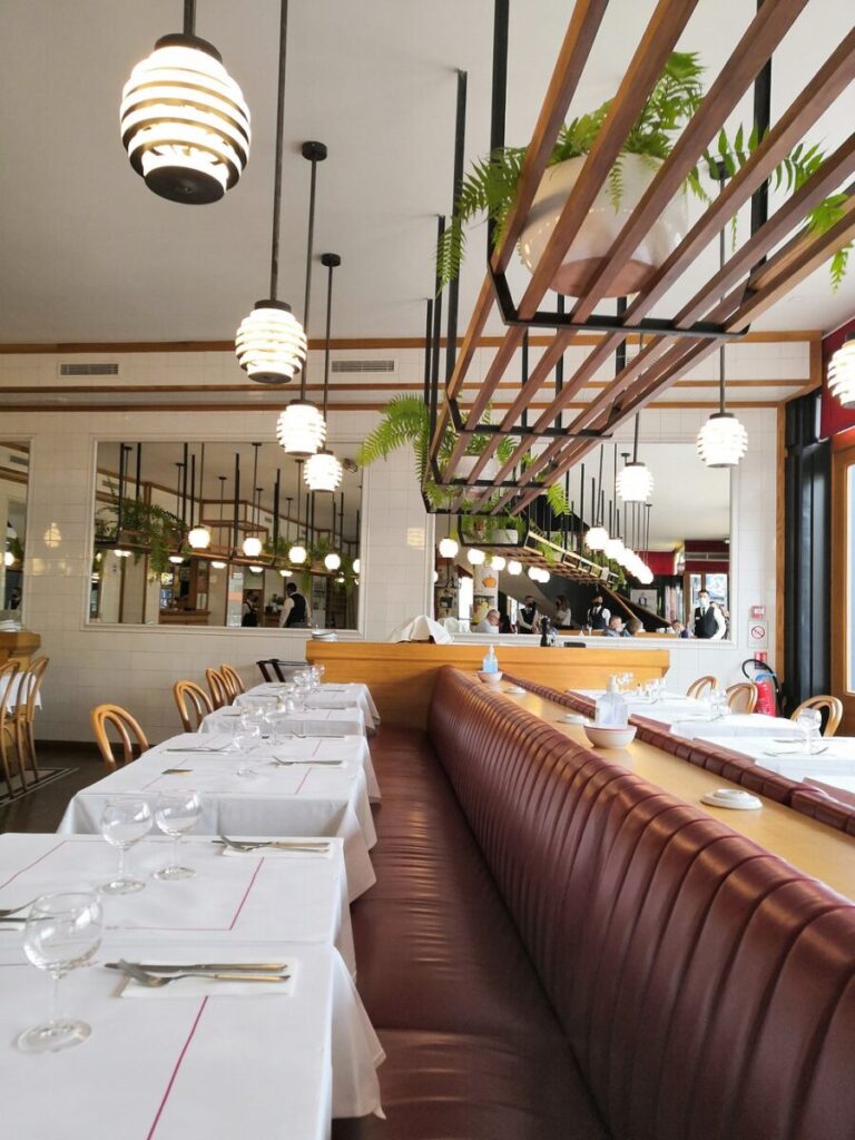 Cozy restaurant interior with hanging lights, lush ferns, wooden accents, and tables set with white linens and glassware, evoking an inviting ambiance.