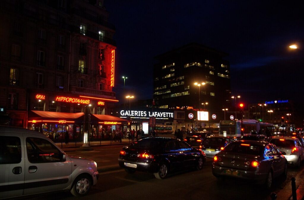 Nighttime scene of cars navigating Boulevard du Montparnasse, surrounded by urban lights and shadows.