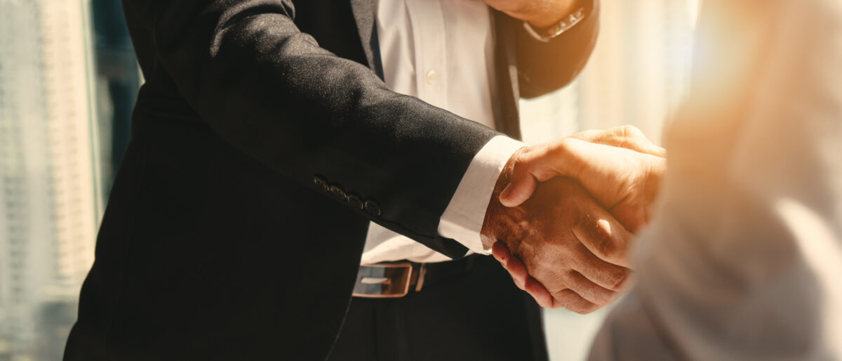 Two people in business attire shake hands in a sunlit office, symbolizing agreement and professionalism. The setting conveys a warm, collaborative atmosphere.