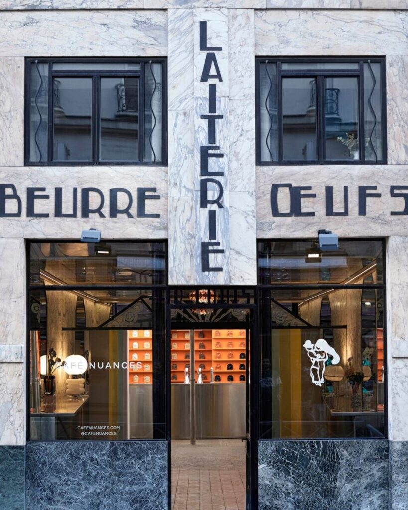 Facade of a stylish Café Nuances cafe with marble and black accents. Large windows show an inviting, modern interior with warm lighting and decorative elements.
