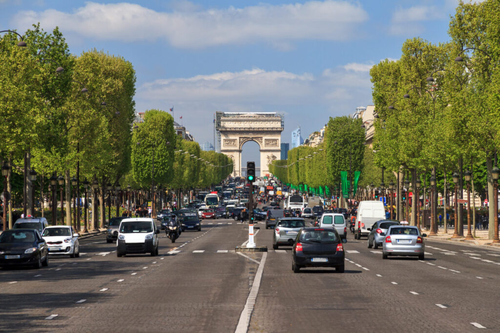 Champs-Élysées with heavy traffic under clear skies. The Arc de Triomphe stands at the end of the tree-lined avenue, creating a lively urban scene.