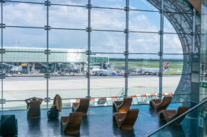 Looking out a large ellipsoid window at Charles De Gaulle airport with two rows of chairs in silhouette in front of it.