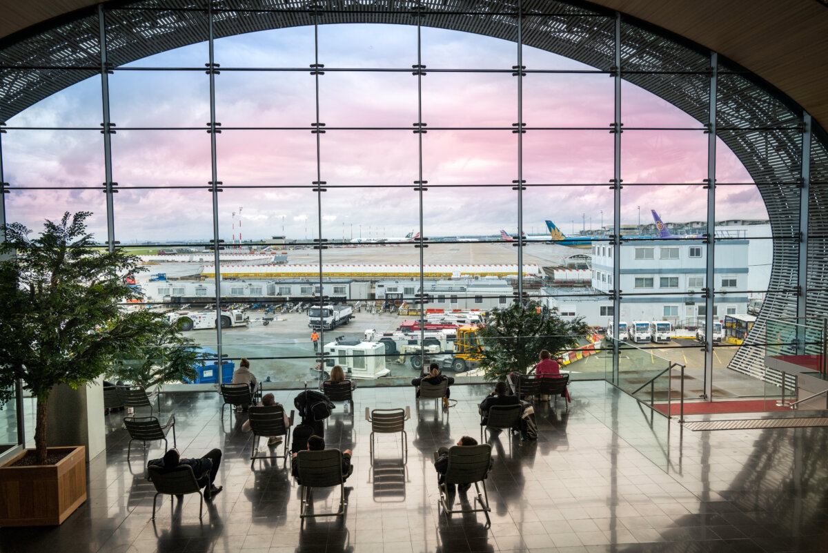 Tourists seated in the departure lounge gaze out at the airfield through a large oval window at Charles de Gaulle Airport.