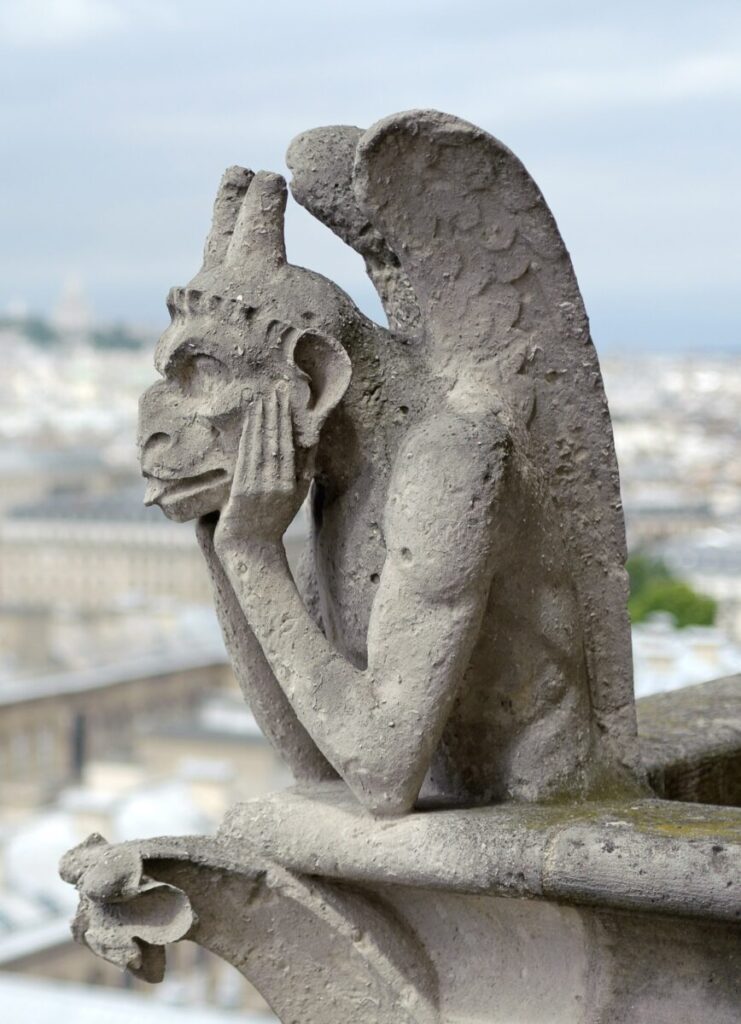 Notre-Dame stone gargoyle perched on a ledge, gazing thoughtfully into the distance. It has wings and rests its chin on its hands, overlooking a blurred cityscape.