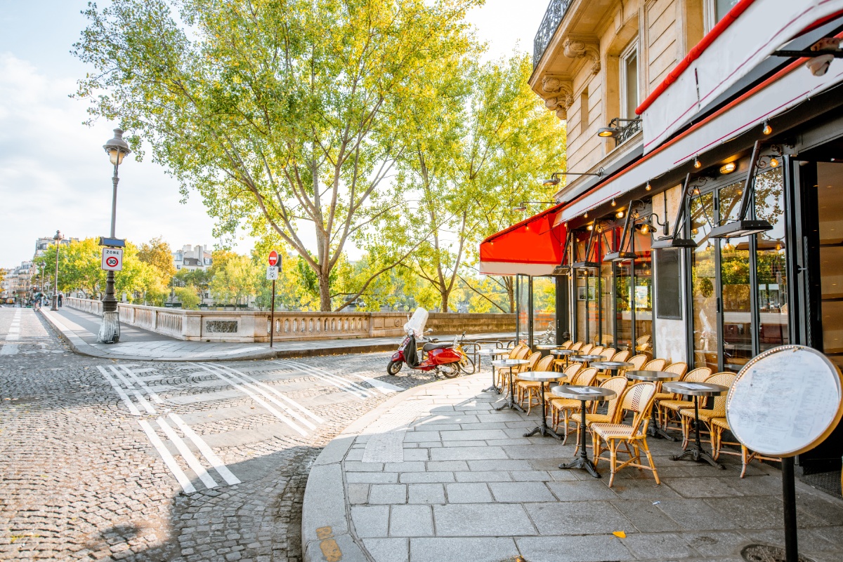 Charming Parisian street corner with a cafe terrace featuring an inviting red awning and outdoor wicker chairs. A red scooter is parked nearby, framed by lush trees.