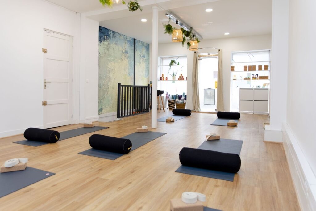 Core Yoga Paris studio with wooden flooring, blue mats, and black bolsters. Soft lighting, plants hanging, and a calming mural create a serene atmosphere.