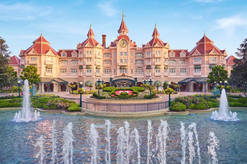 Disneyland Hotel grand, pink fairytale hotel with red spires and a central clock tower, surrounded by gardens and fountains, under a clear blue sky.