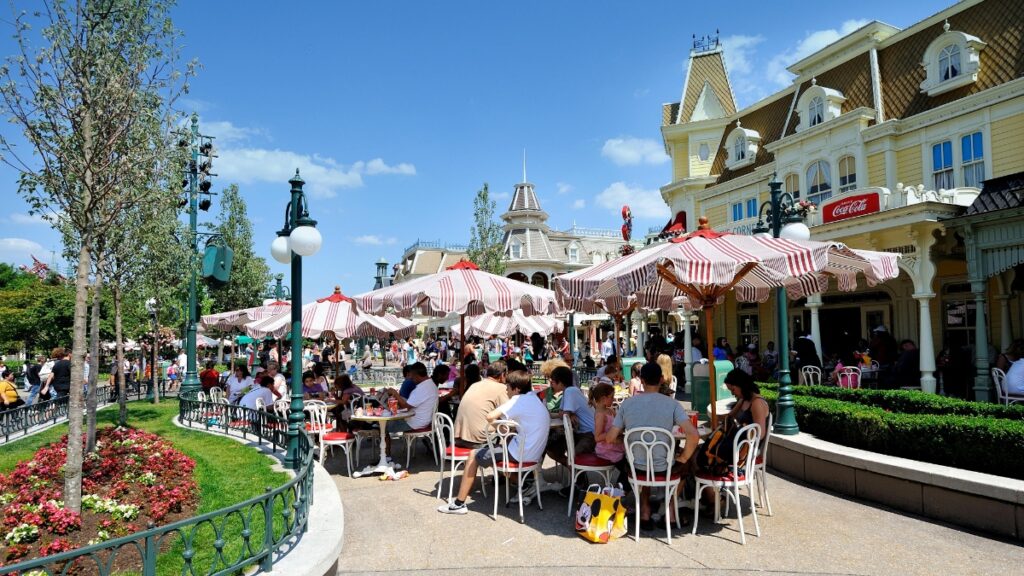 People enjoy dining outdoors at tables with red-and-white striped umbrellas in Casey’s Corner at Disneyland Paris.