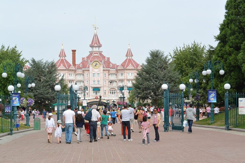 Crowds walk towards a grand, pink castle with towers and a clock. Green iron gates and trees flank the path, creating an inviting, cheerful atmosphere.