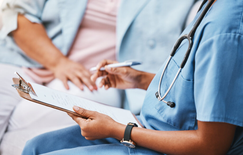Healthcare professional in blue scrubs writes on a clipboard while seated, with a stethoscope draped around their neck. A patient sits nearby, hands on lap.