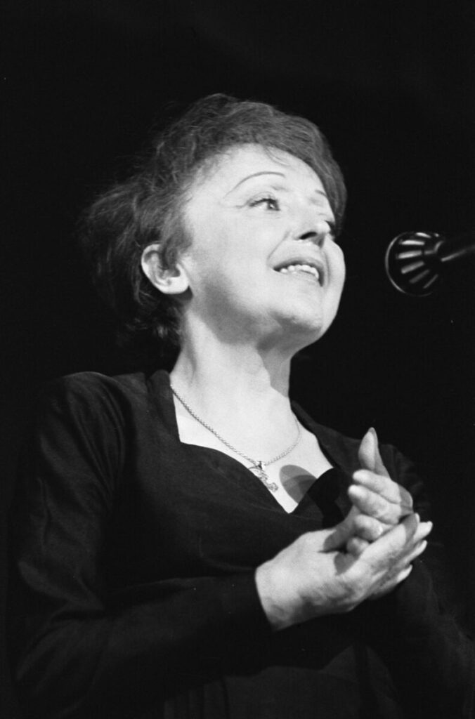 Black and white image of a Édith Piaf speaking into a microphone on stage