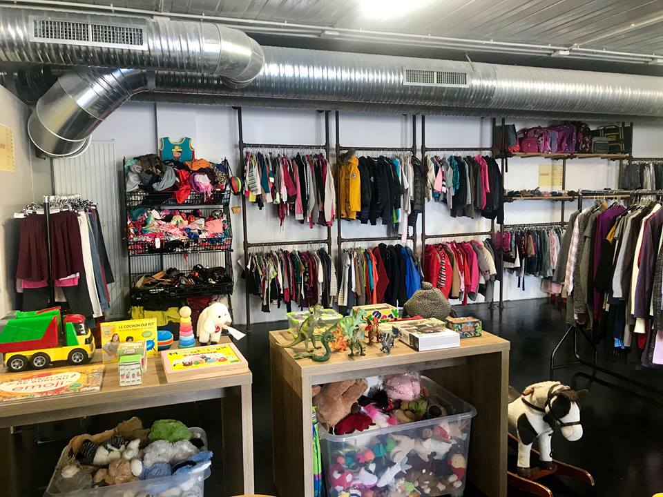 Emmaüs Alternative thrift store with racks of colorful clothing, bins of plush toys, and a toy section with dinosaurs and a model truck. Bright and organized.