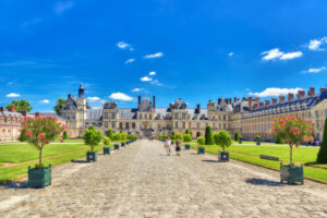Fontainebleau, France grand palace under a bright blue sky with a cobblestone path lined by manicured trees and colorful flowers. People stroll down the path, evoking a serene atmosphere.