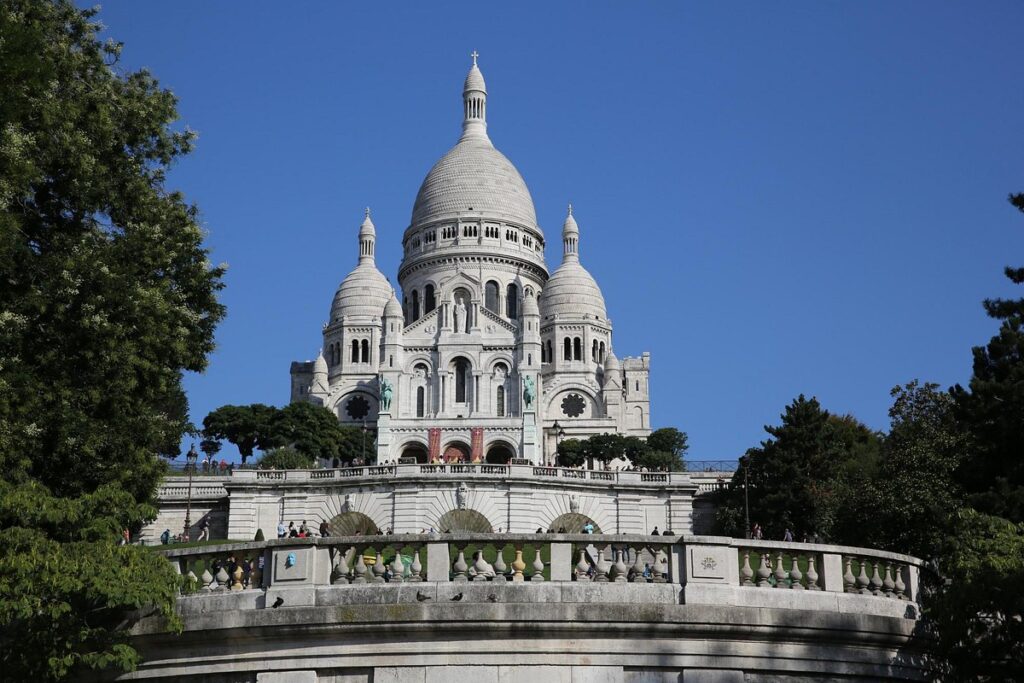 The iconic Montmartre Funicular ascends towards the Basilica of the Sacré-Cœur, visible against a clear blue sky, with tourists on the surrounding terraces.