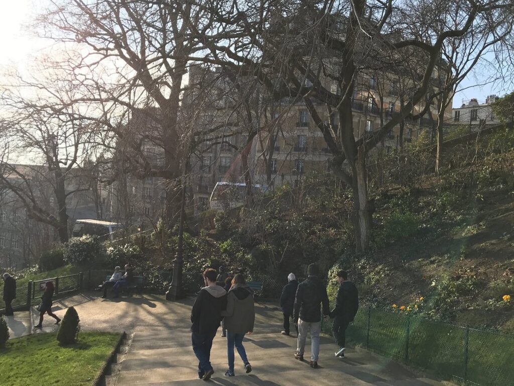 A serene pathway in Montmartre leading towards the Funicular, lined with trees and benches, with people walking and enjoying the calm environment.