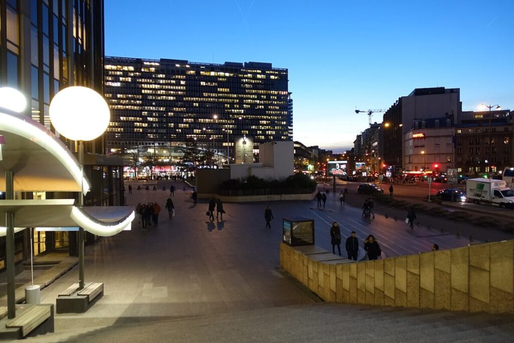 Night view of a city square with the prominent Gare de Paris-Montparnasse building illuminated in the background.
