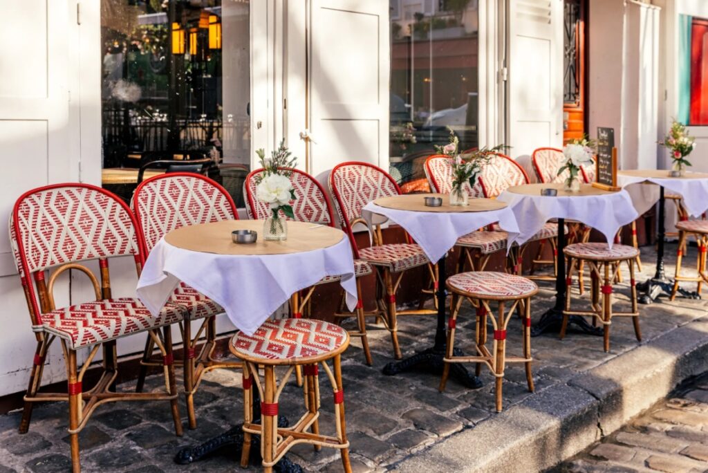 Outdoor café scene of Grace Café with round tables, white tablecloths, and red-patterned chairs on cobblestone. Flowers and vases add charm and elegance.
