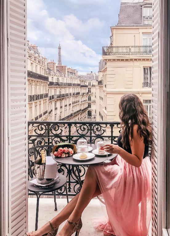 A woman in a pink dress enjoys a leisurely breakfast on a balcony with a view of Parisian rooftops and the Eiffel Tower in the distance.