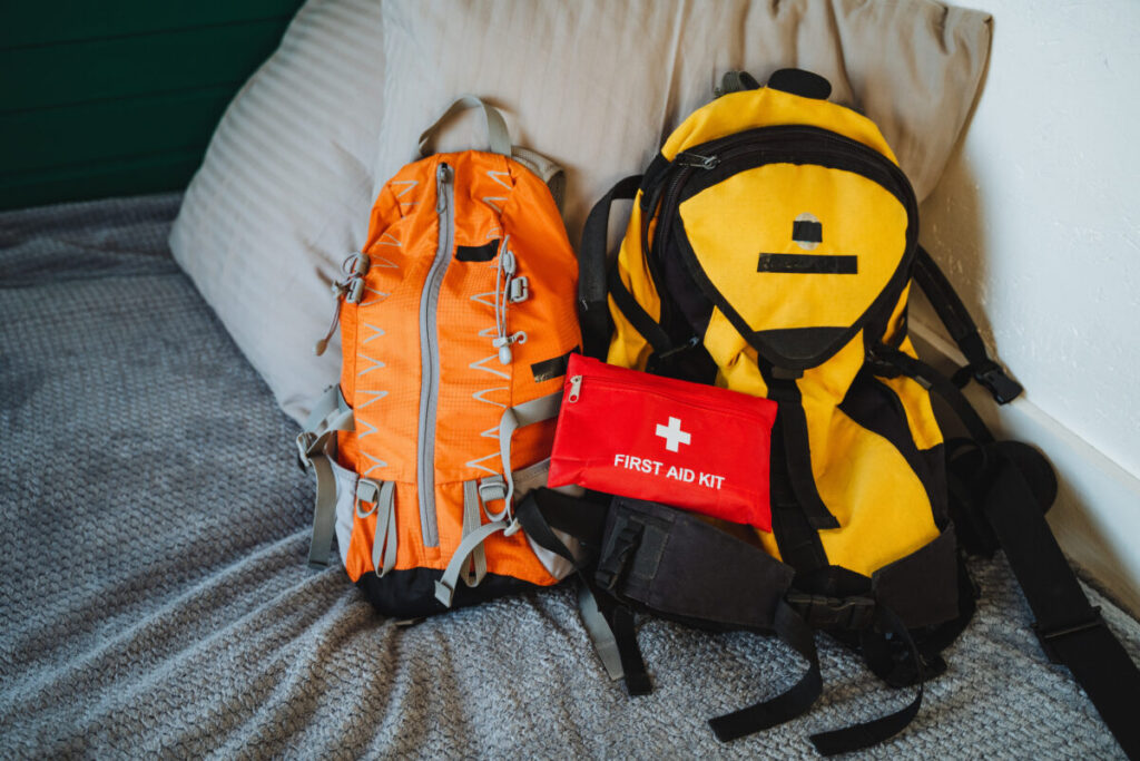 Two backpacks, one bright yellow, sit on a cozy bed next to a neatly arranged first aid kit, ready for outdoor adventures.