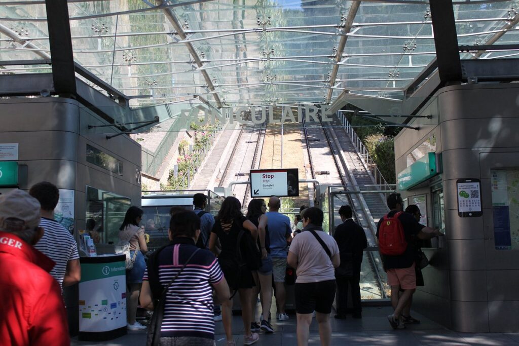 Inside the Montmartre Funicular station, passengers wait in line at the ticket barrier with the Funicular's inclined tracks visible through the glass structure ahead.