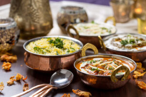 Copper bowls of Indian cuisine on a wooden table, featuring colorful curry, rice, and fragrant garnishes. The scene is warm and inviting.