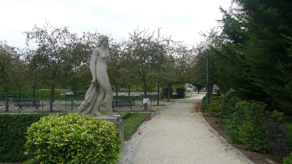 In Jardin de Reuilly, Paris, a statue of a woman is located on a path adjacent to a park bench.