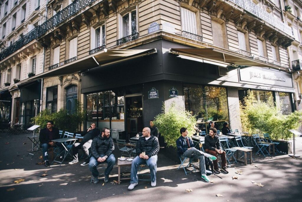 Group of people sits outsideKB CaféShop a charming Parisian café on a sunny day. The mood is relaxed, with classic architecture and lush plants adding a cozy ambiance.