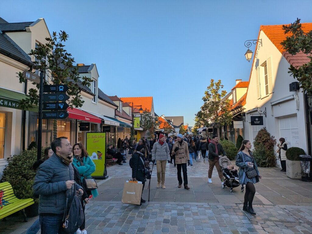 People stroll along a bustling street in La Vallée Village, France, surrounded by shops and fellow visitors.