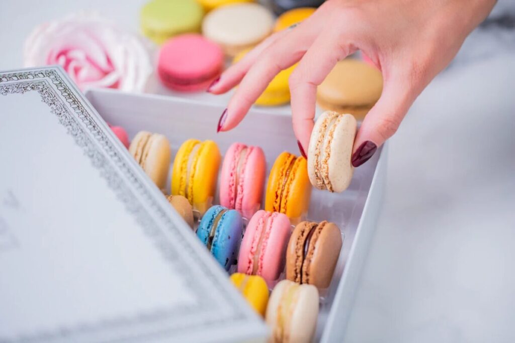 Hand with dark red nails selects a white macaron from a box filled with colorful macarons, including yellow, pink, and blue, on a white surface.