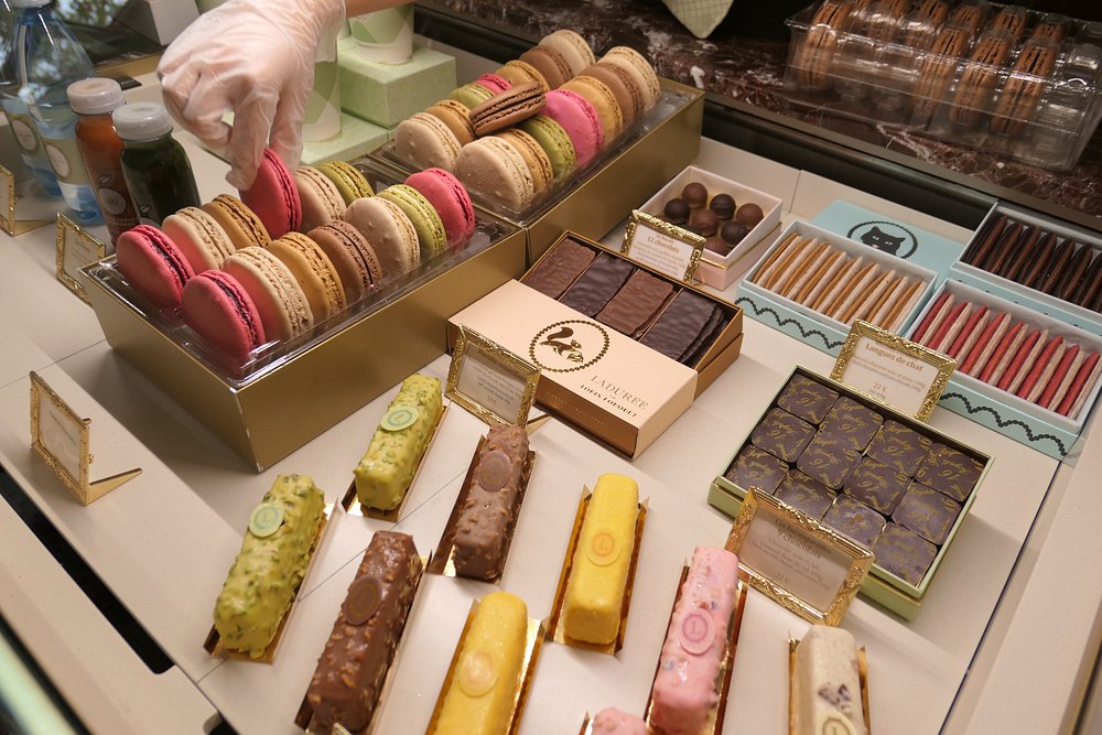 Ladurée, Paris display showcasing colorful macarons, assorted pastries, and chocolates. A gloved hand selects a pink macaron.