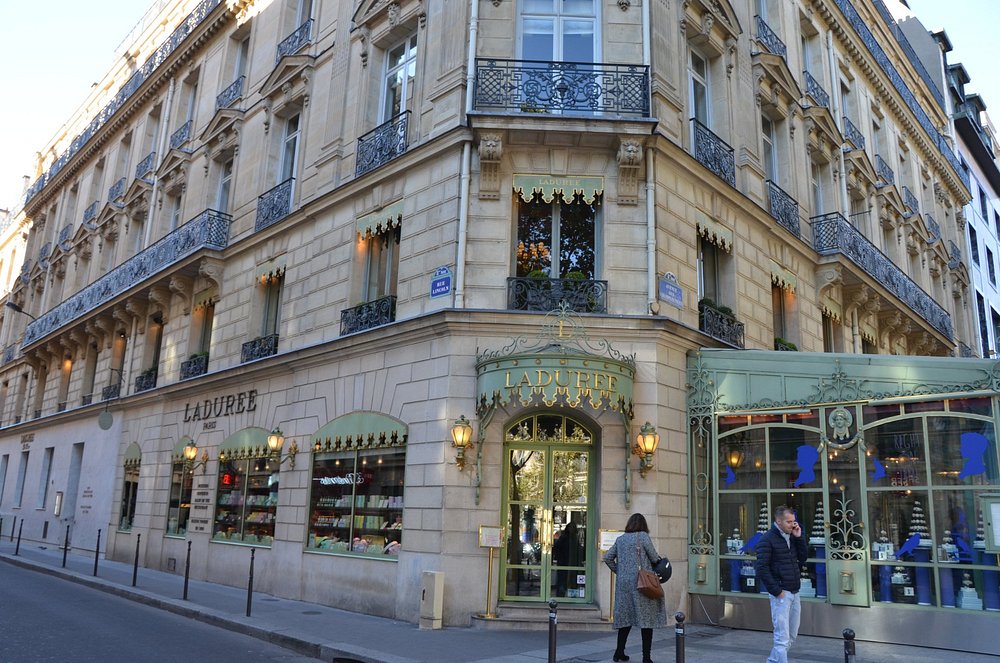 Parisian street scene with an elegant, ornate corner building housing Ladurée. Its light green storefront features decorative windows and cheerful passerby.
