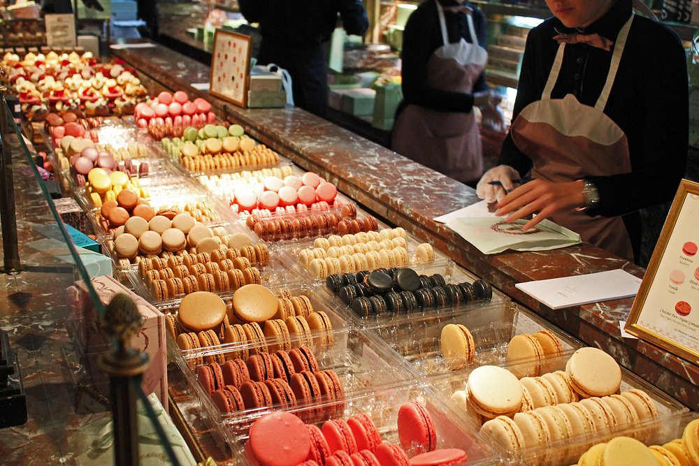 Ladurée, Paris, France vibrant display of assorted macarons in various colors on a marble counter, with a person in an apron arranging pastries, creating a luxurious, inviting atmosphere.