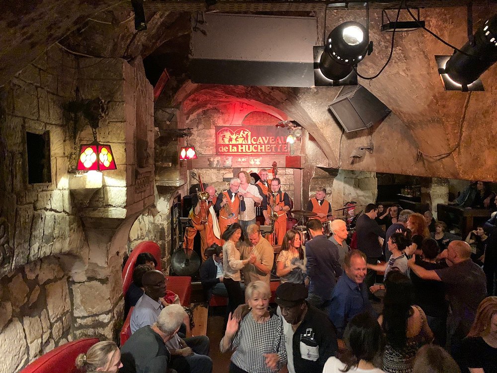 Inside Le Caveau de la Huchette in Paris, a bustling crowd mingles, standing and enjoying drinks in a lively atmosphere.
