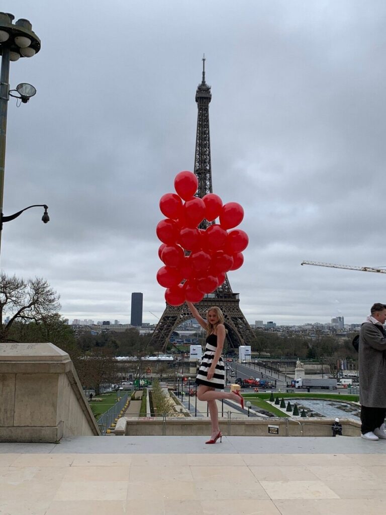 A woman holding a cluster of red balloons stands in front of the Eiffel Tower on an overcast day.