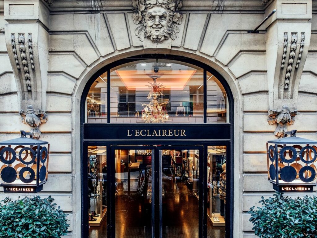Elegant storefront of L'Eclaireur with an ornate stone façade, featuring a sculpted face, decorative lights, and large glass doors. Luxurious ambiance.